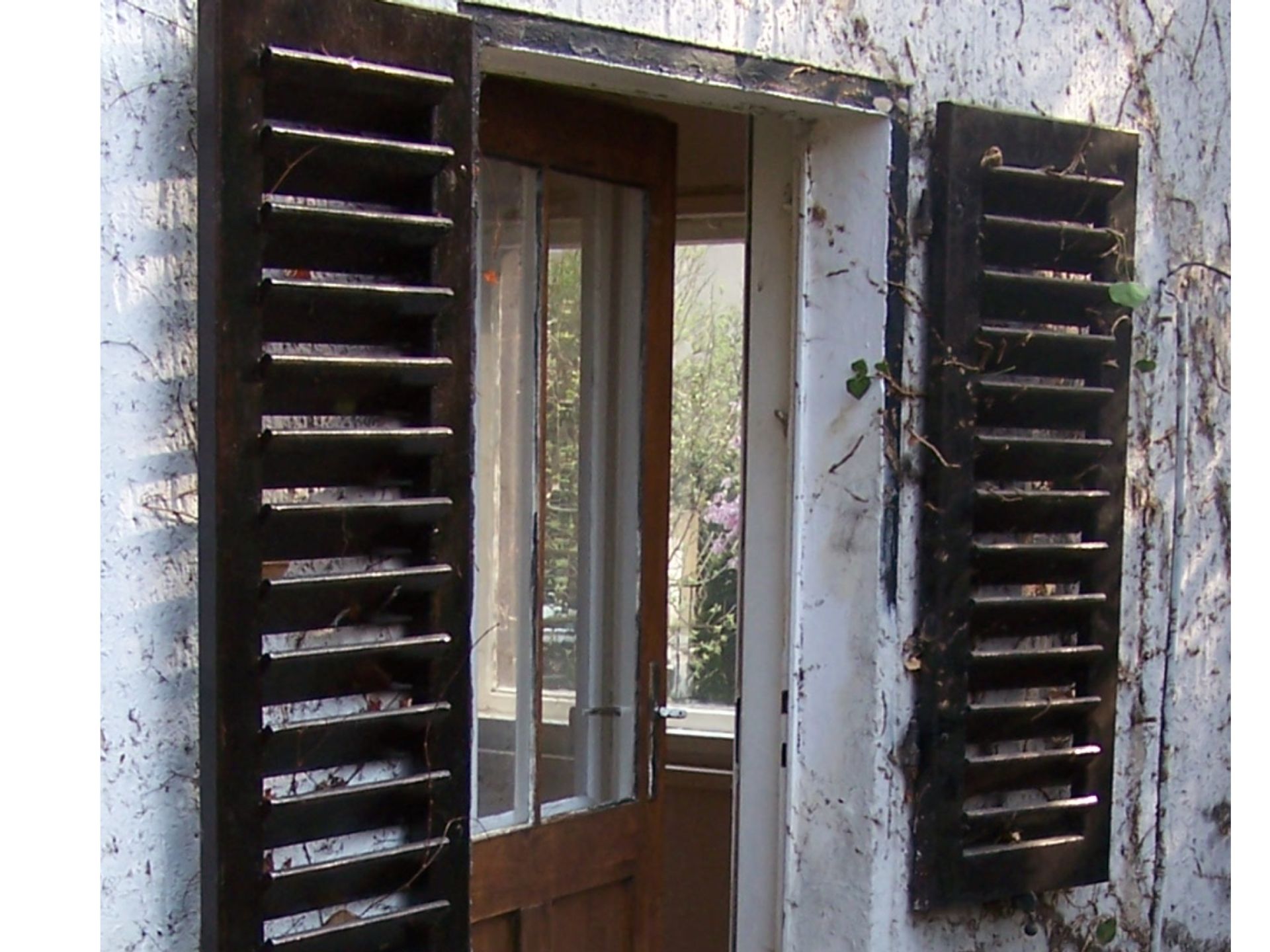 Braune Fensterläden rahmen eine offene Tür mit weißem Vorhang an einer weißen, strukturierten Wand ein.