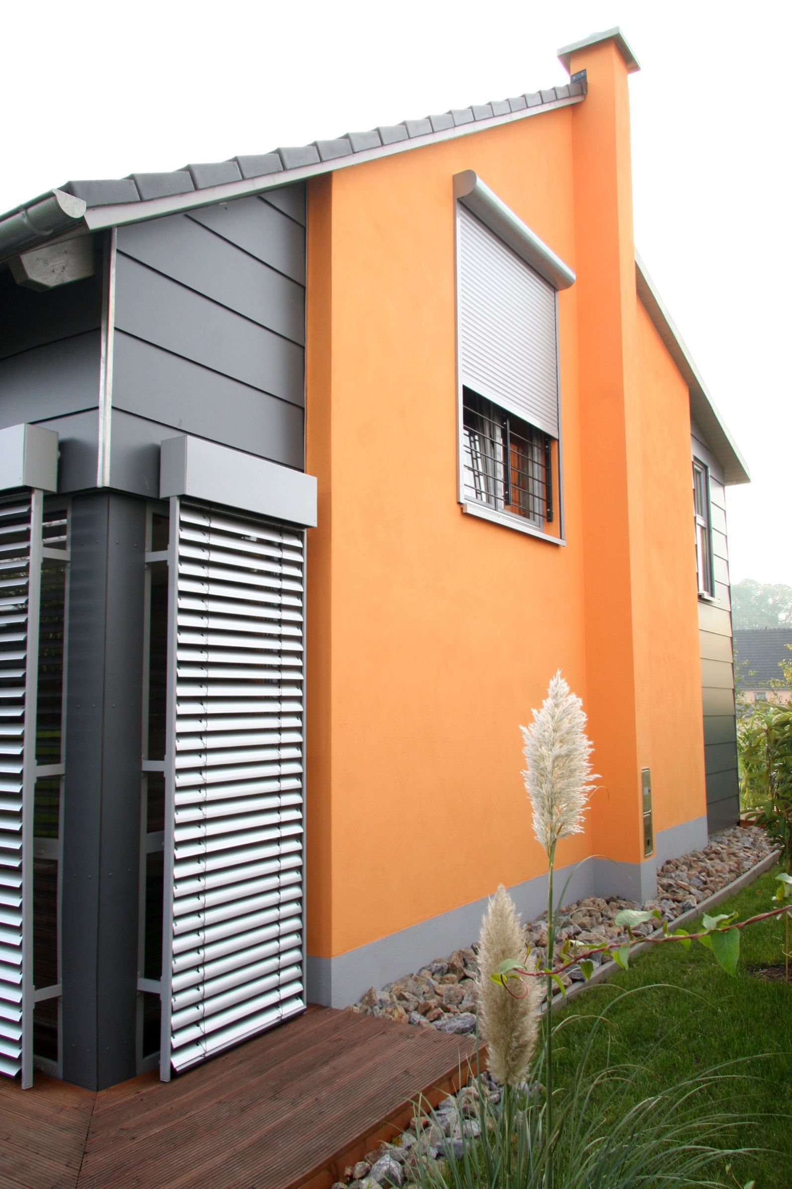 Orangefarbenes Haus mit grauen Akzenten; Fenster mit Fensterladen, Schornstein. Holzterrasse und Rasen.