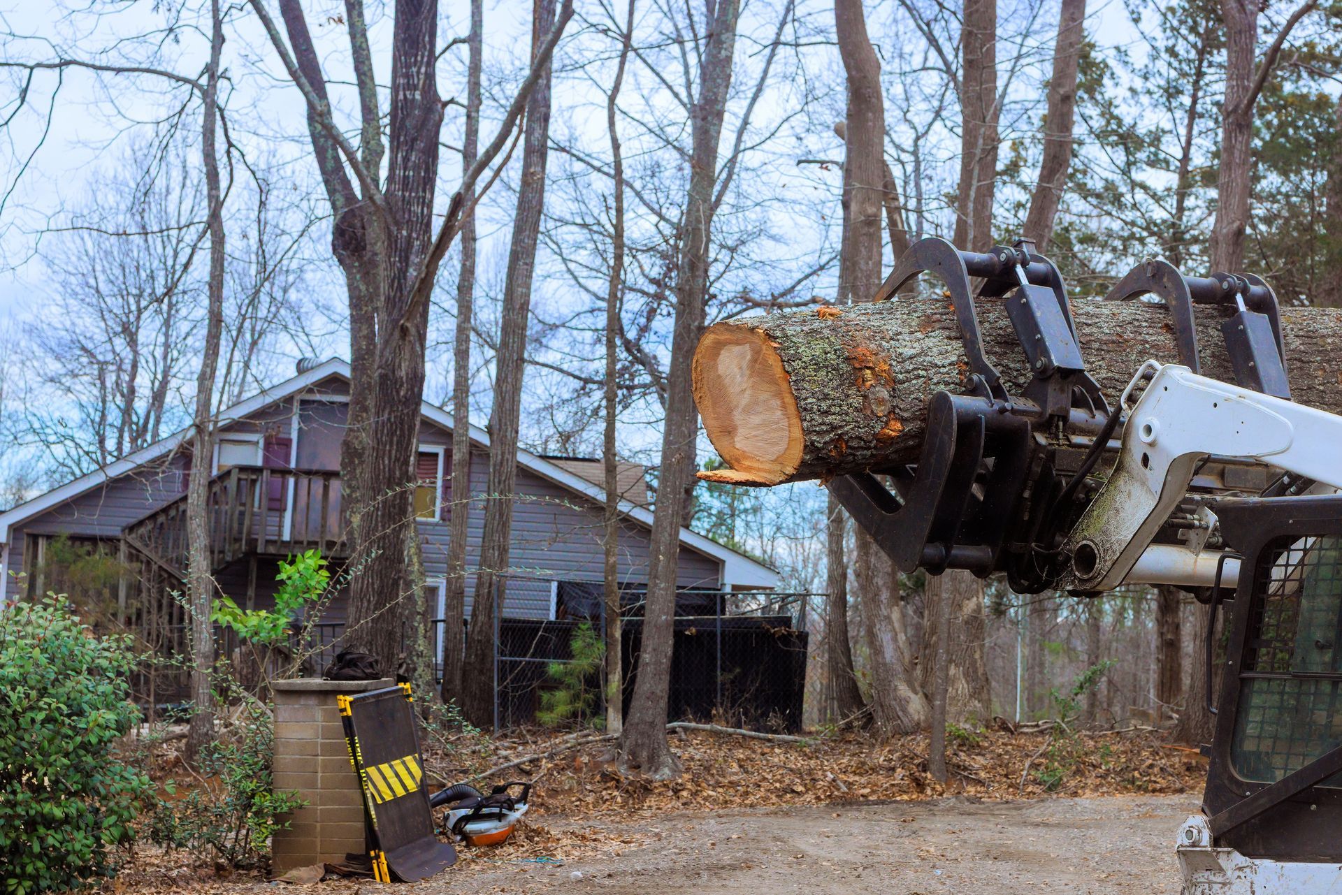 Backhoe lifting a large log beside a house in a wooded yard