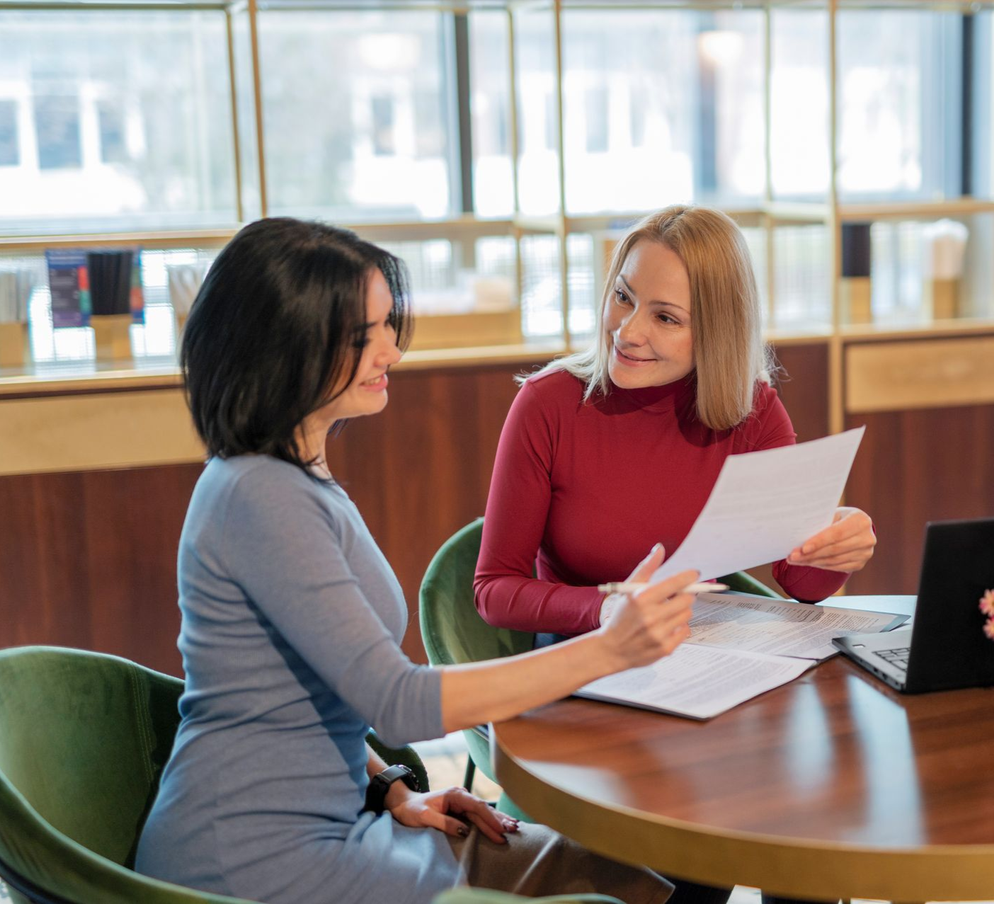 Two women discussing papers at a round table in a bright office lounge