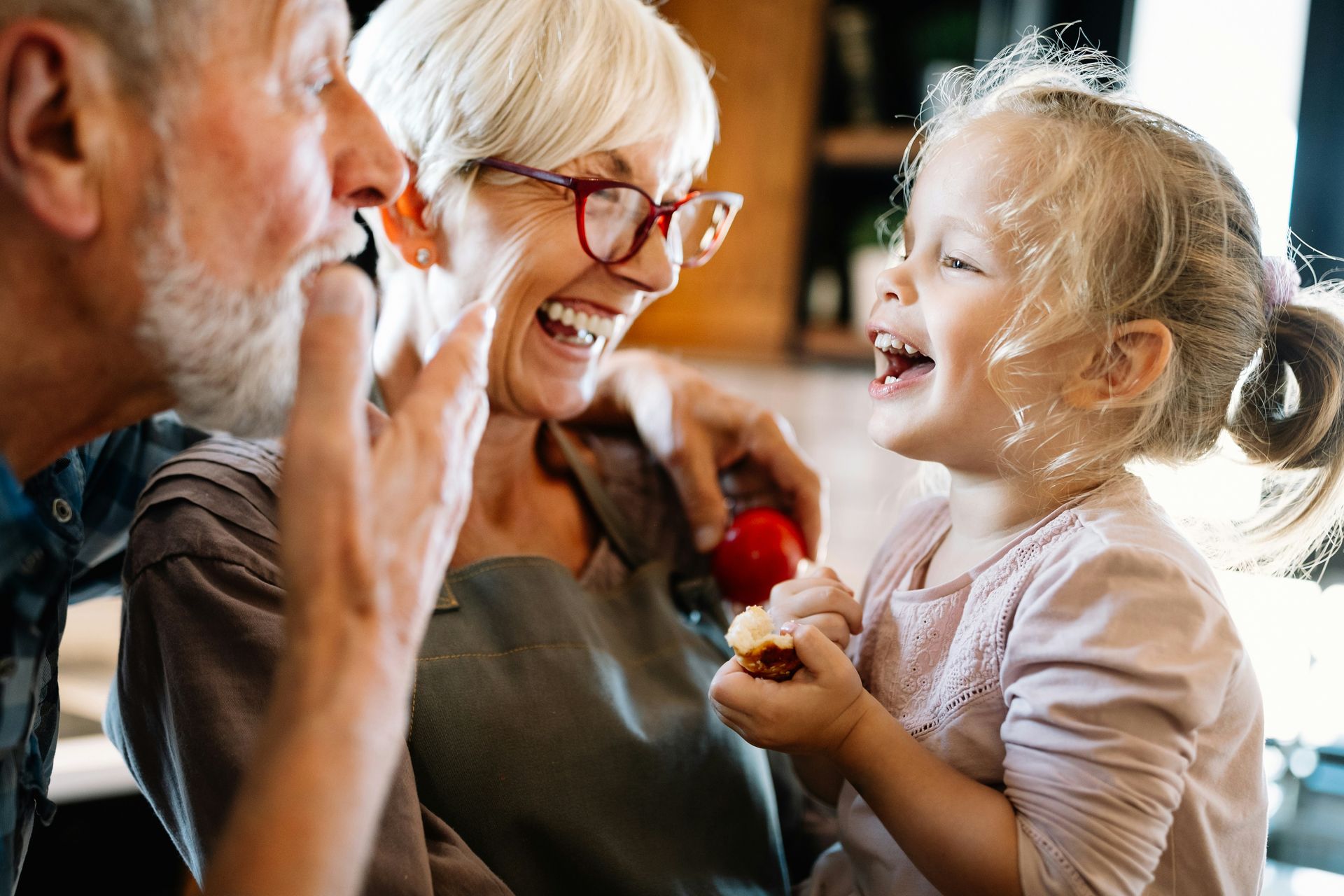 Three people laughing together indoors, with a child holding a red flower.