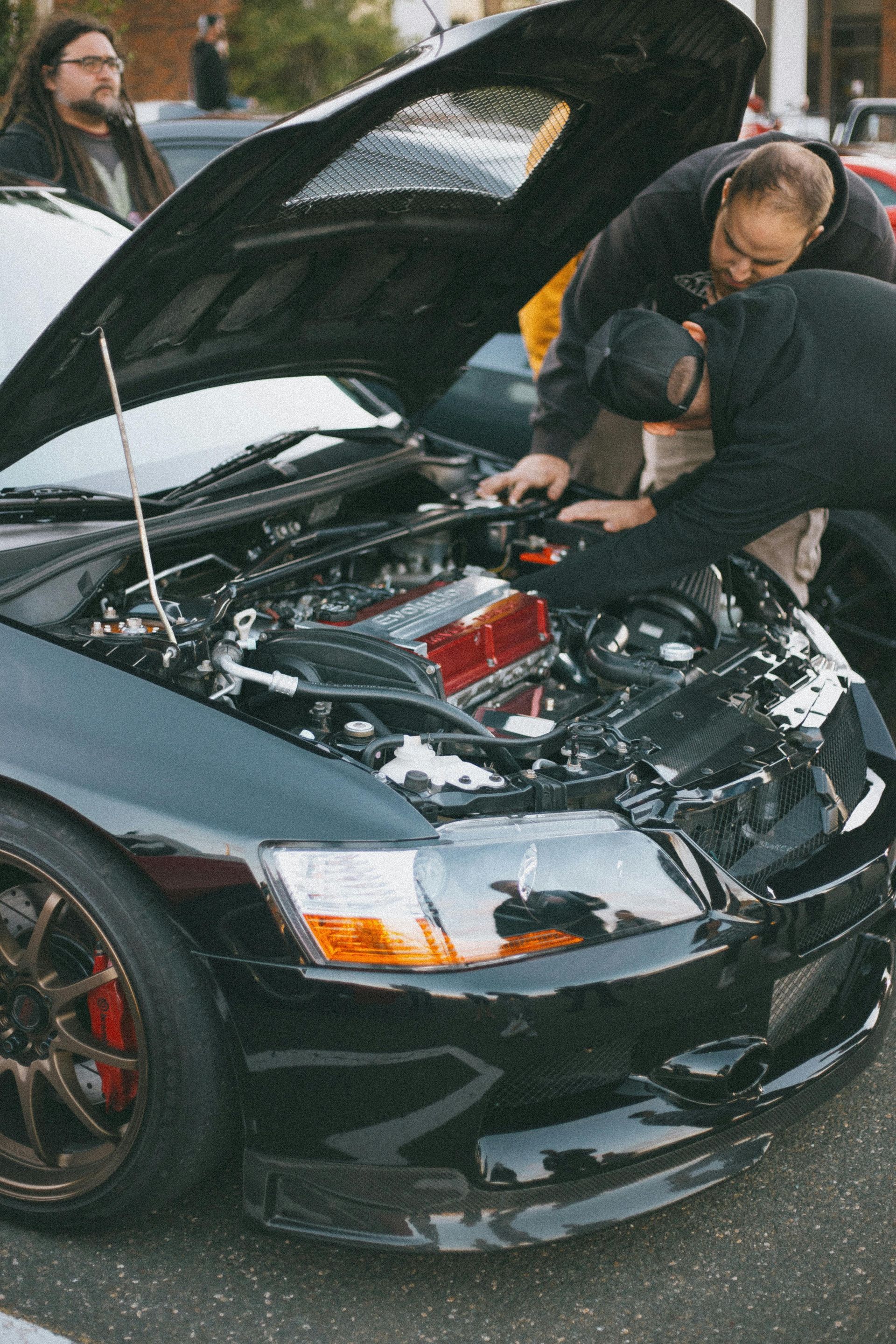 Three people inspect a black car's open engine bay at an outdoor event.