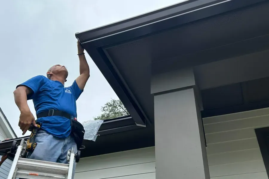 A man is standing on a ladder fixing a gutter on the side of a house.