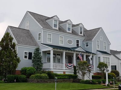 Large gray suburban house with white trim, multiple gables, and a front porch with patriotic bunting and flags