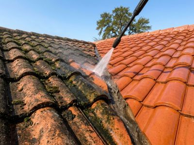 Pressure washer cleaning a red clay roof beside a dirt-streaked black roof under a blue sky
