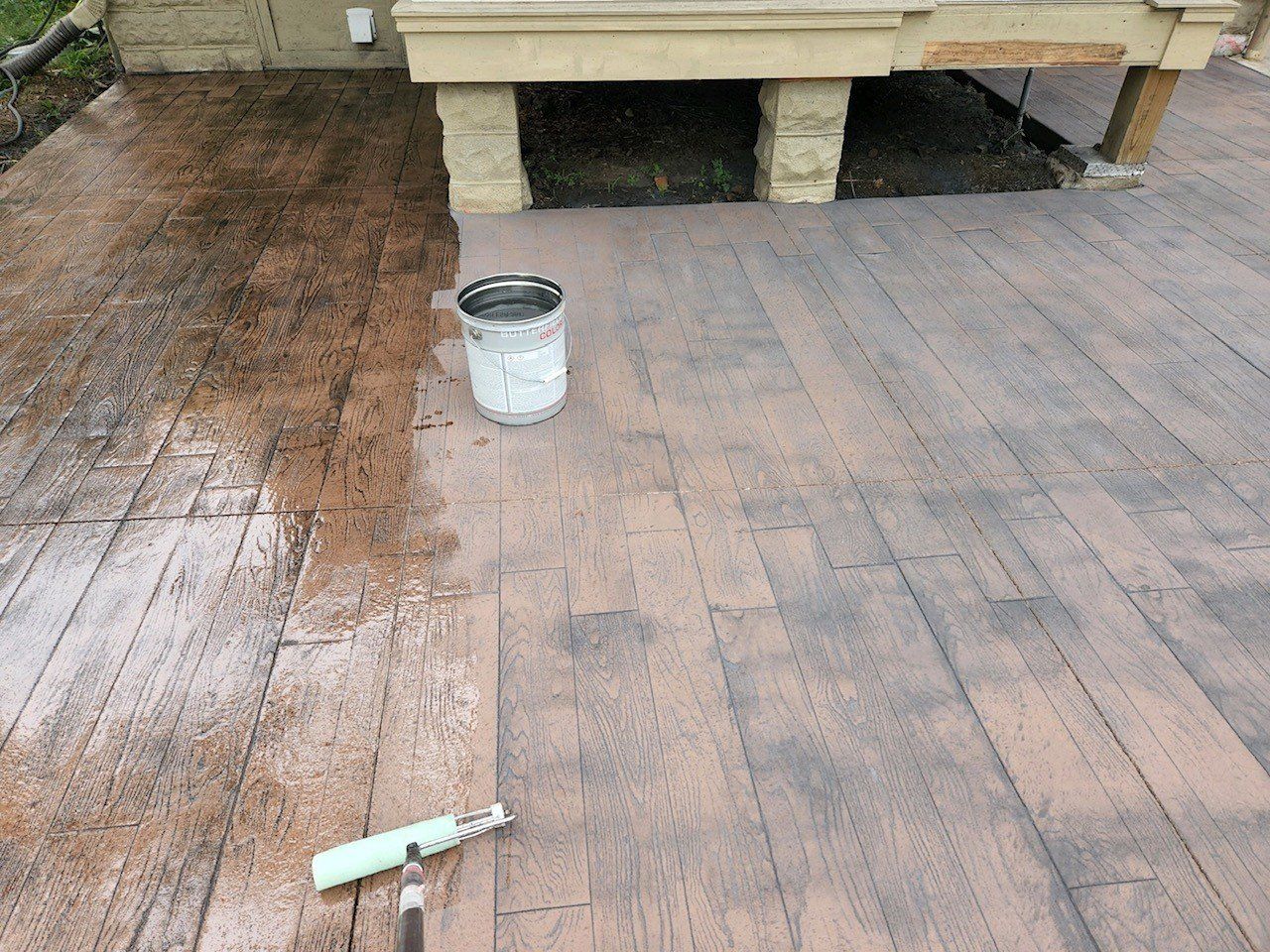 Applying stain to a wooden deck with a roller; a bucket of stain sits nearby.