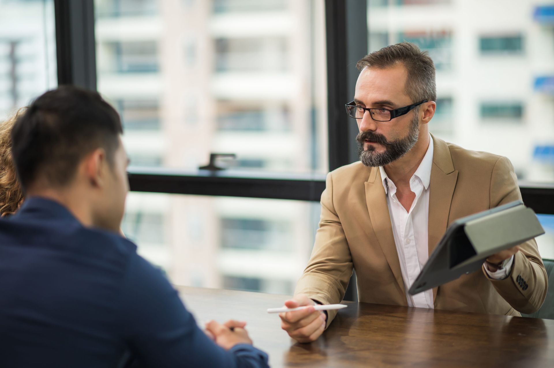 Two people in a business meeting, one holding a tablet and speaking across a table.