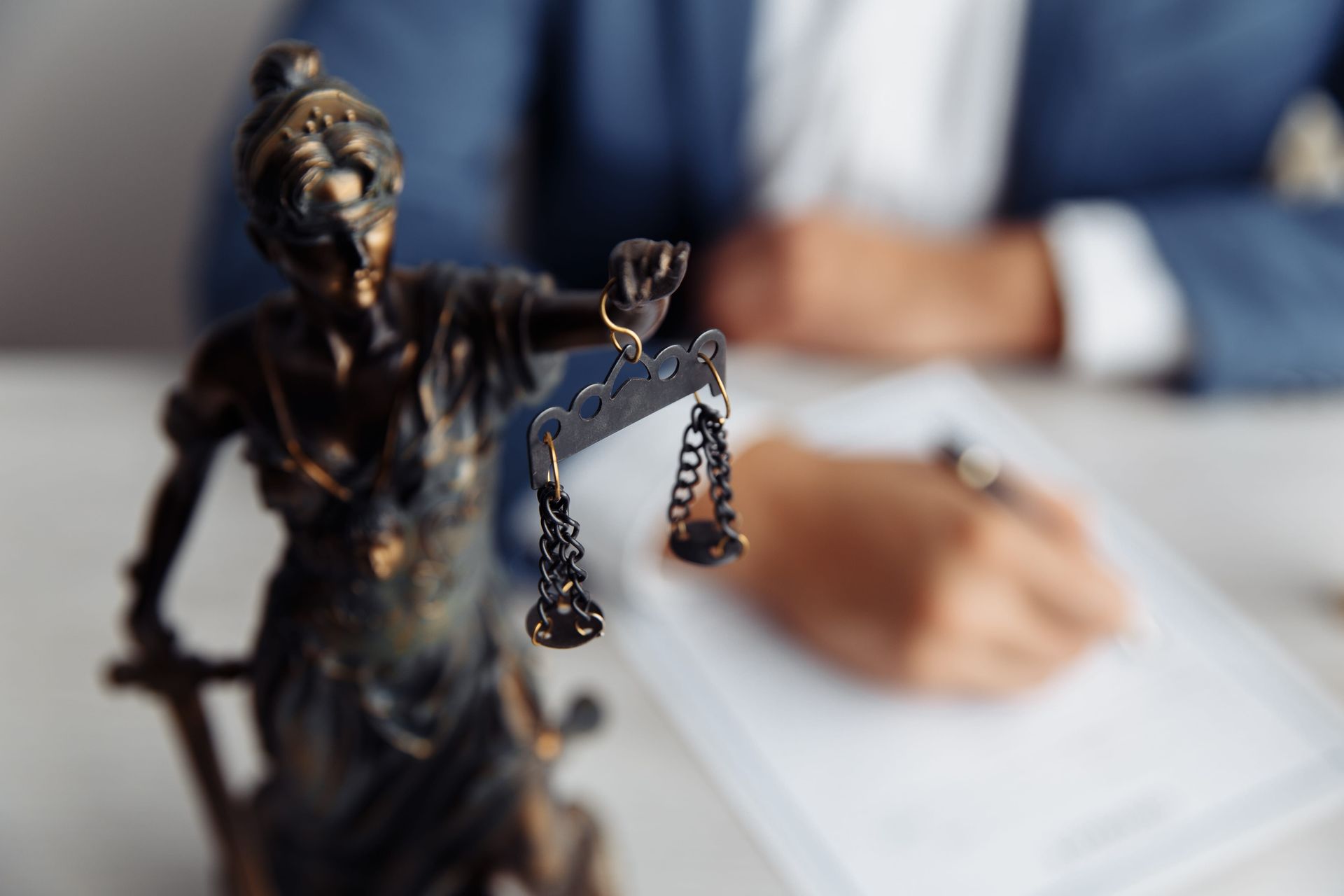 Lady Justice statue with scales in foreground, blurred person writing on paper in background
