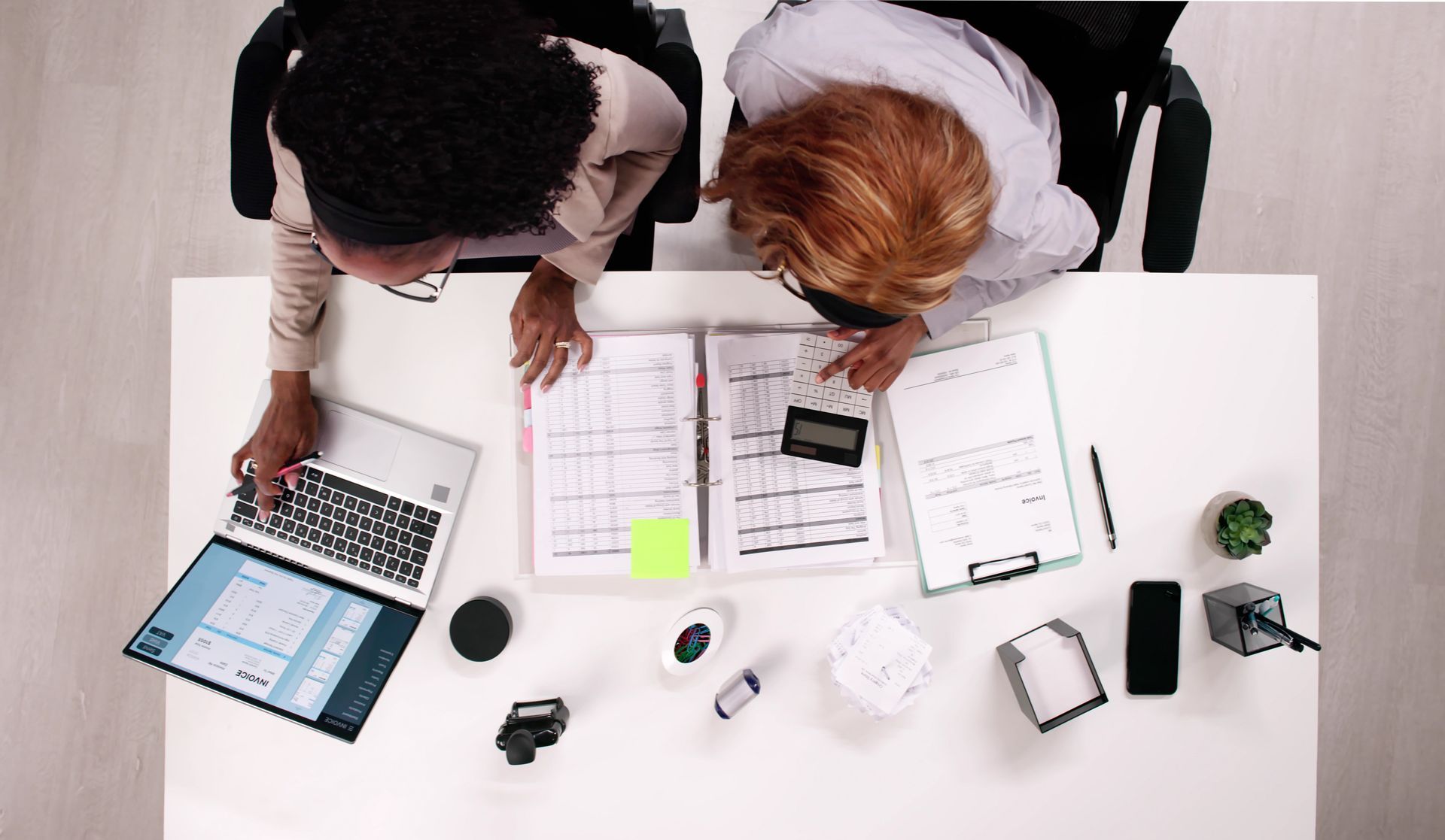 Two people at a white desk, working. One uses a laptop; both examine papers and a calculator.