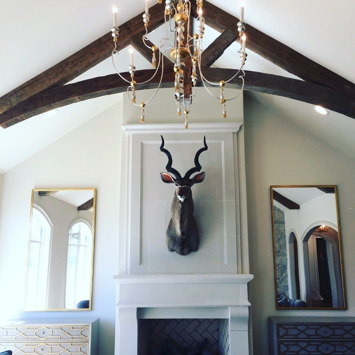 White fireplace with mounted antelope head beneath dark beams in a vaulted room, flanked by mirrors and a chandelier
