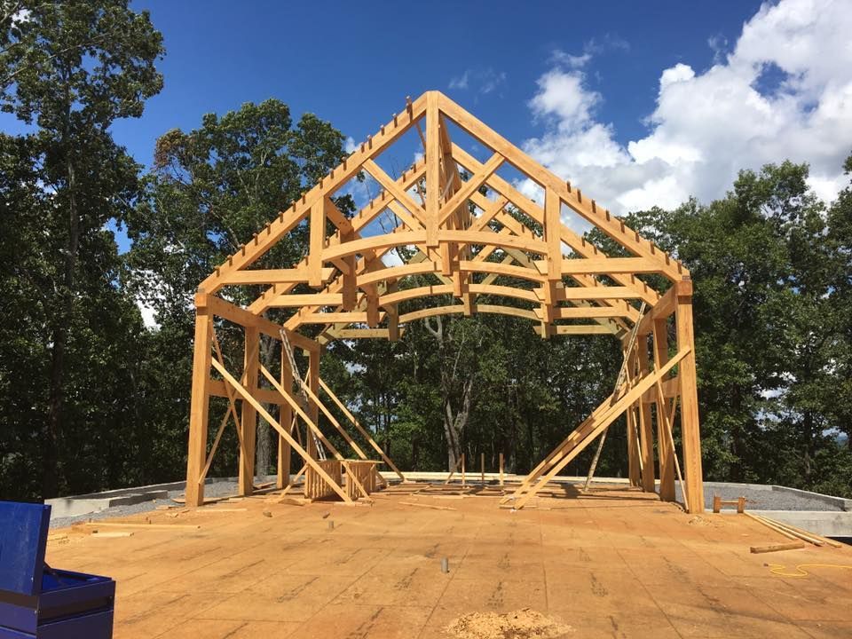 Wooden house frame under construction on a cleared lot, with blue sky and trees in the background