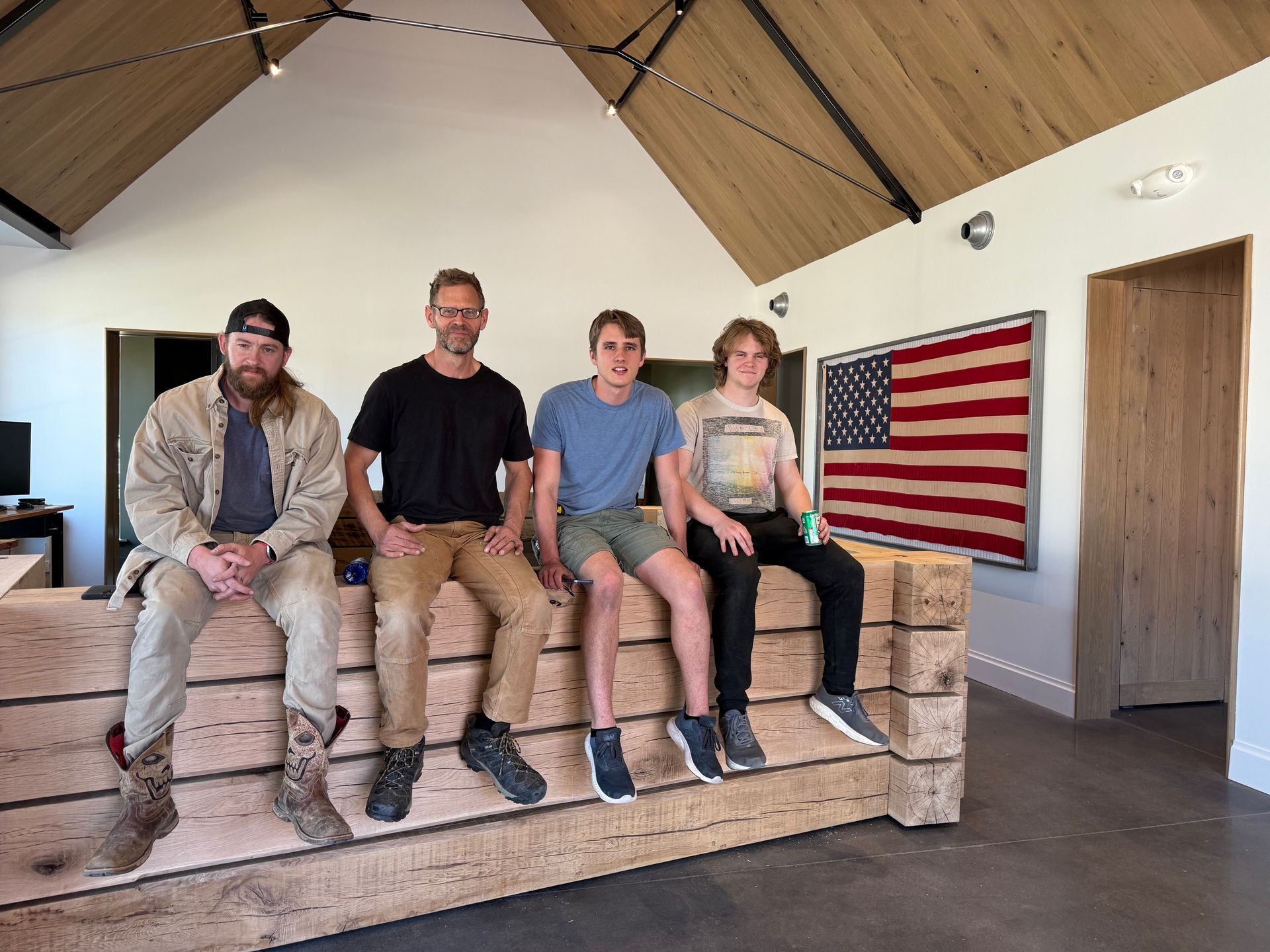 Four people sit on a wooden bench in a bright room beside a large American flag.