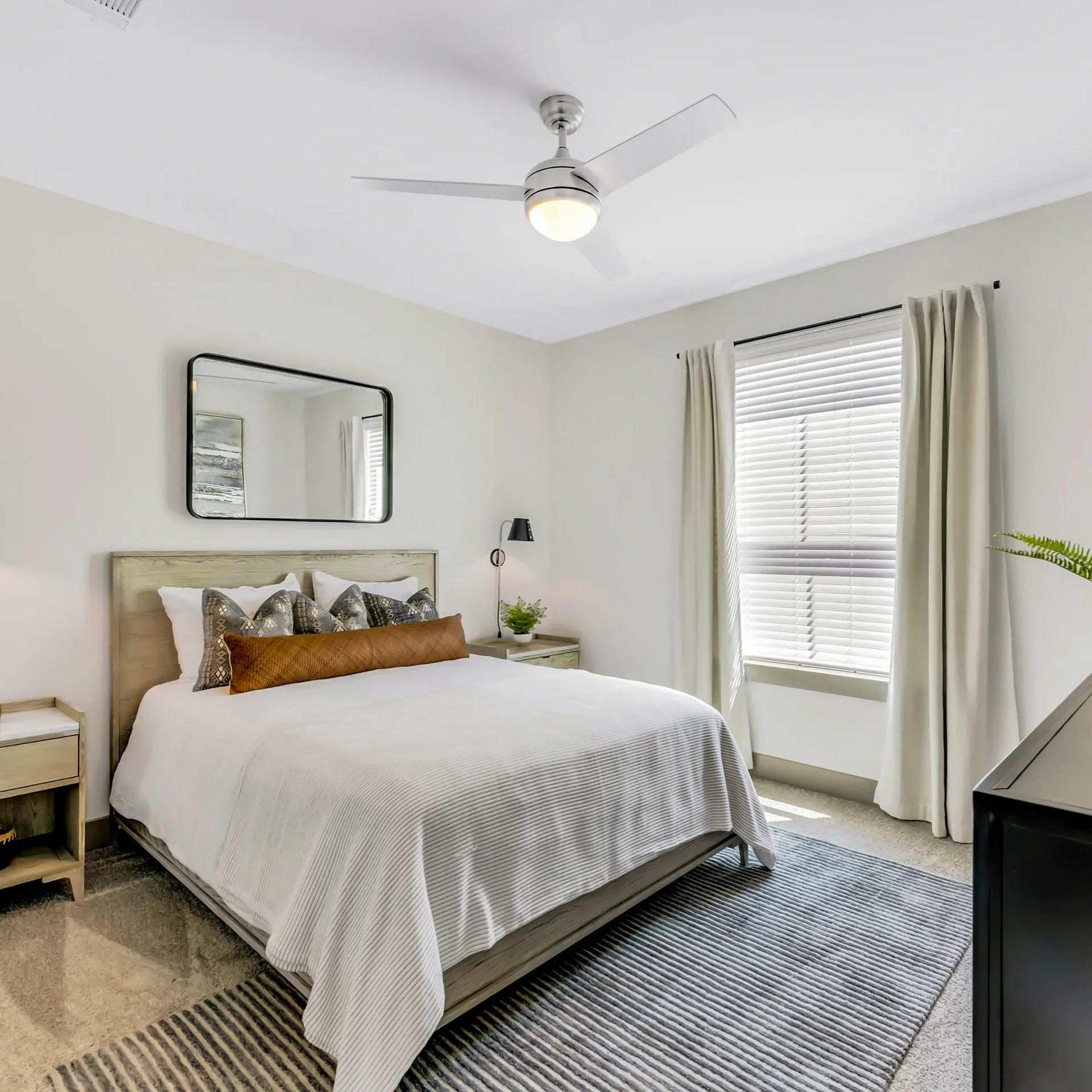 Bedroom with bed, mirror, window, and ceiling fan. Gray and white color scheme at Atlas Bluewood Apartments in Celina, Texas