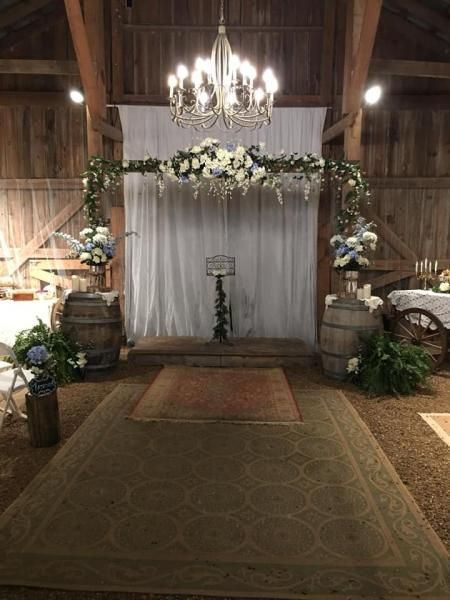 Rustic wedding ceremony arch with white drapery, floral arrangements, chandelier, and aisle runner in a barn venue