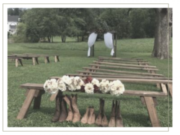 Rustic outdoor ceremony setup with benches and white floral arrangements in a grassy park, two white dresses hanging in back