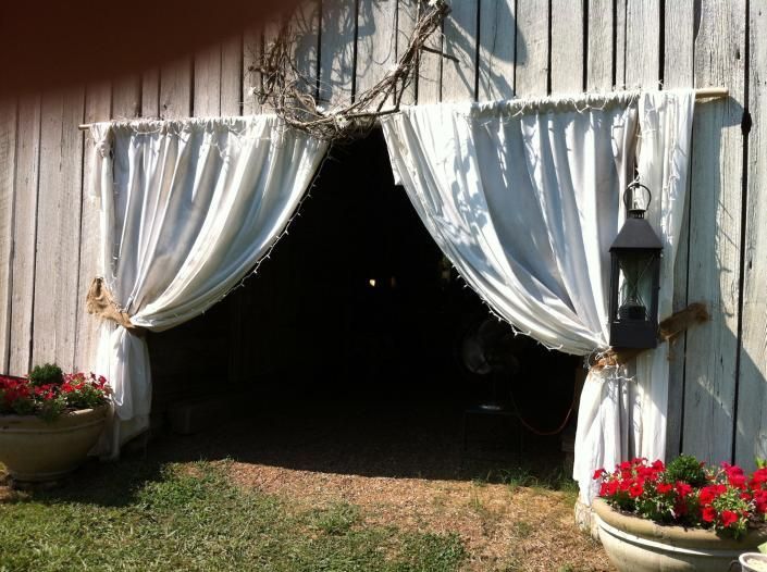 White-draped barn entrance with red flower pots and a hanging lantern, opening into a dark interior