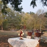Outdoor table with white lace cloth and flower centerpiece in a garden, with chairs and trees in the background