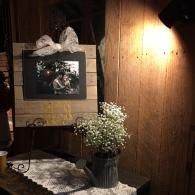 Rustic wooden wall with framed photo, lit wall sconce, lace cloth, and vase of white flowers on a table