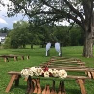 Outdoor wedding setup with wooden benches, floral arrangement, and two people in light blue near an oak tree.