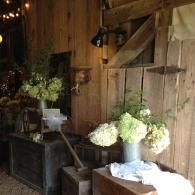 Rustic barn interior with white flower arrangements on tables against a wooden wall.