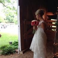 Bride in white dress holding pink bouquet, standing in a barn doorway with greenery outside