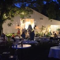 Outdoor evening wedding reception in a lit tent with guests seated at round tables under trees.