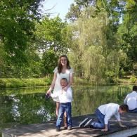 Family on a wooden dock by a pond in a green park, with children playing near the water