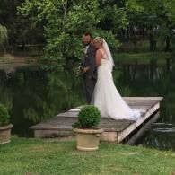 Bride and groom standing on a wooden dock by a calm pond surrounded by trees