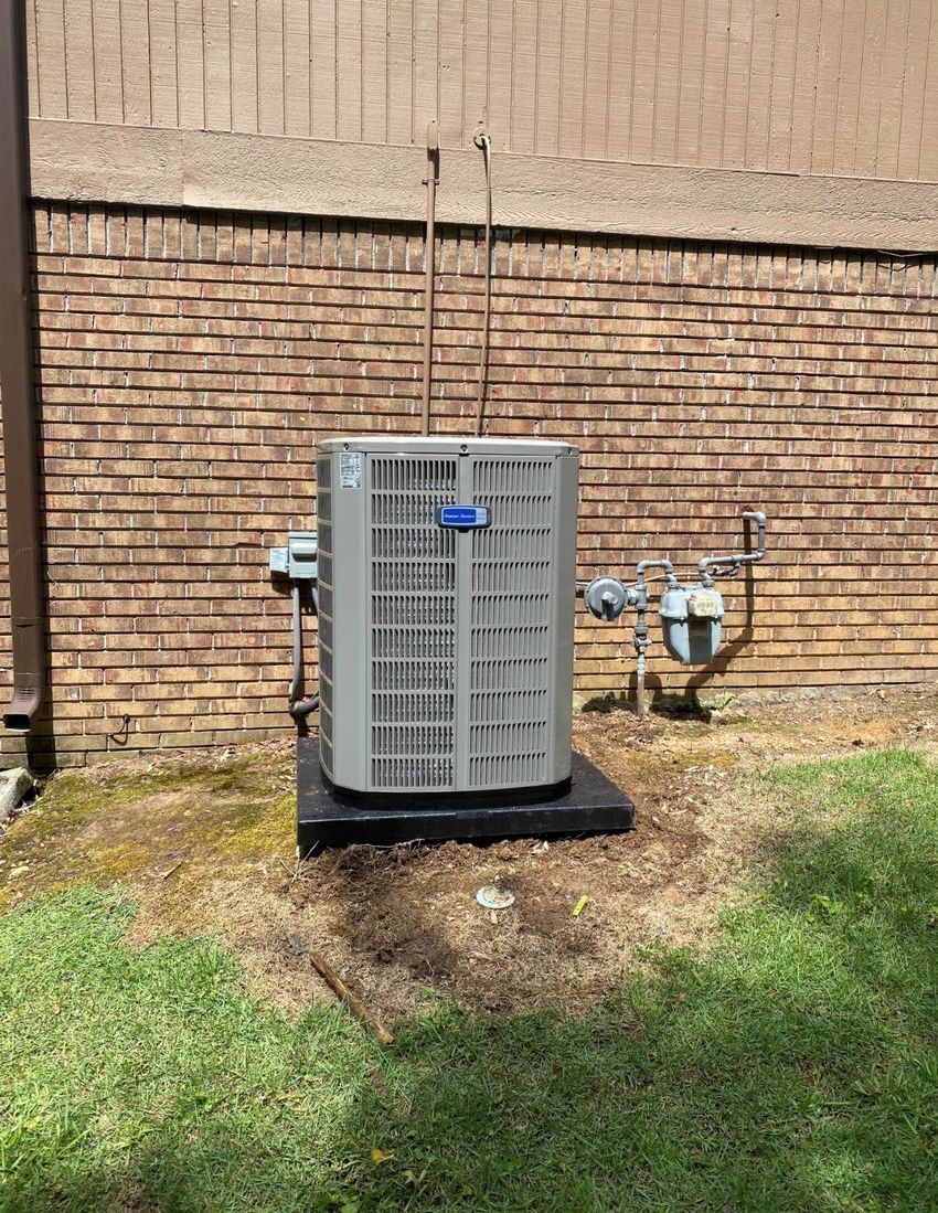 Outdoor HVAC condenser unit against a brick wall, surrounded by grass and dirt.