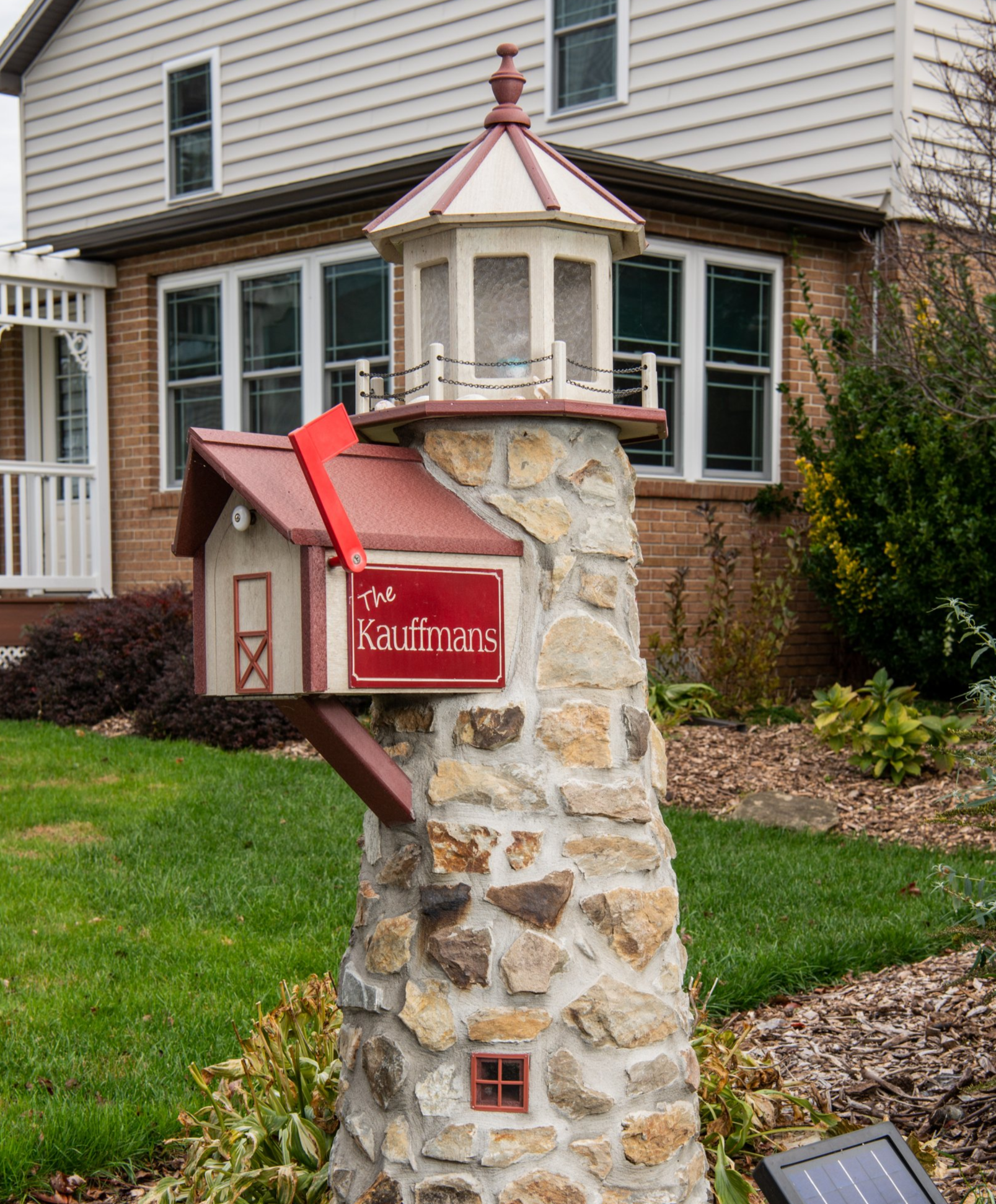 Lighthouse-shaped mailbox made of stone, with a red mailbox attached. A house is in the background.