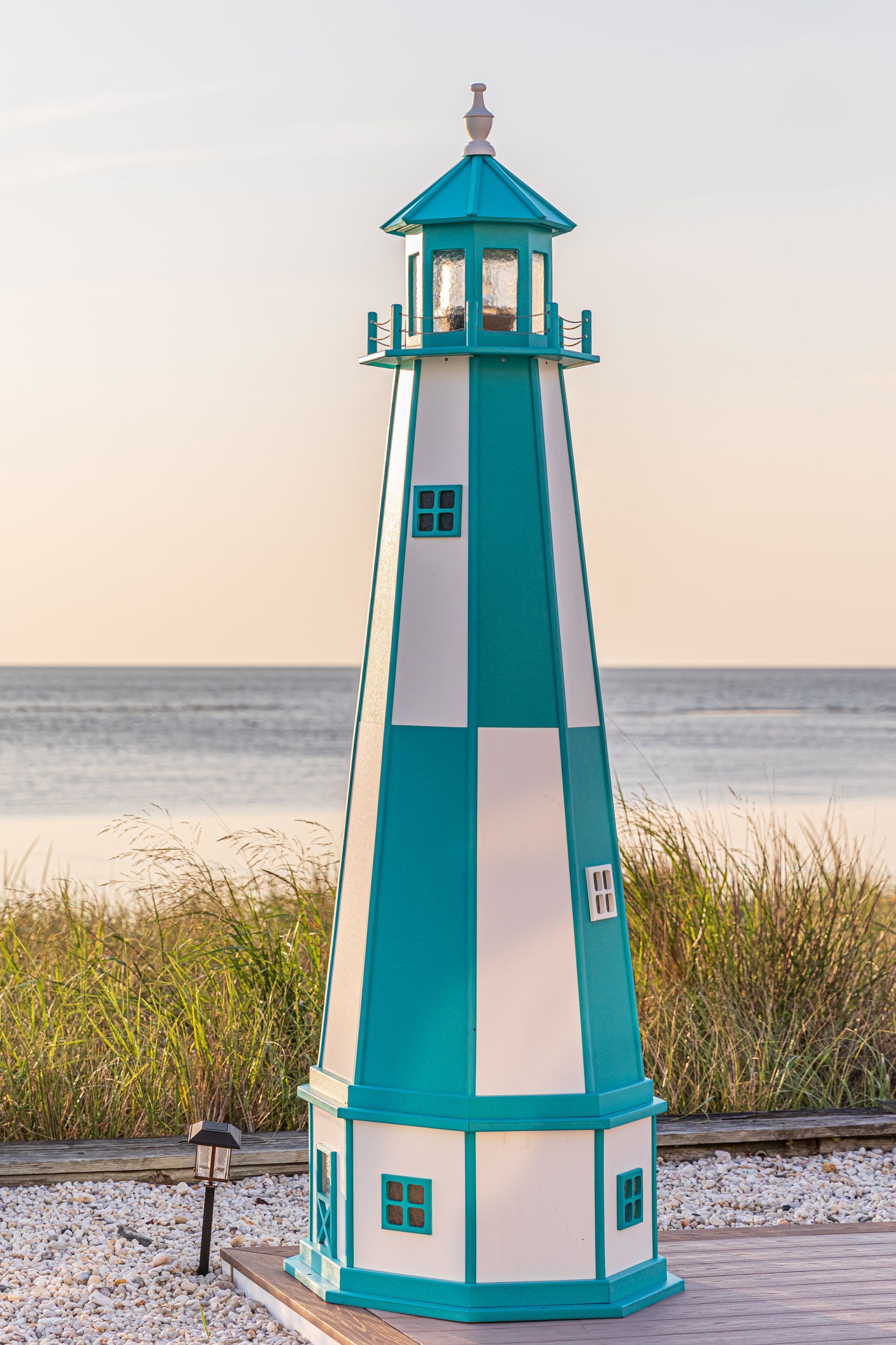 A teal and white decorative lighthouse model stands outdoors near a shoreline with tall grass and a pebble ground.