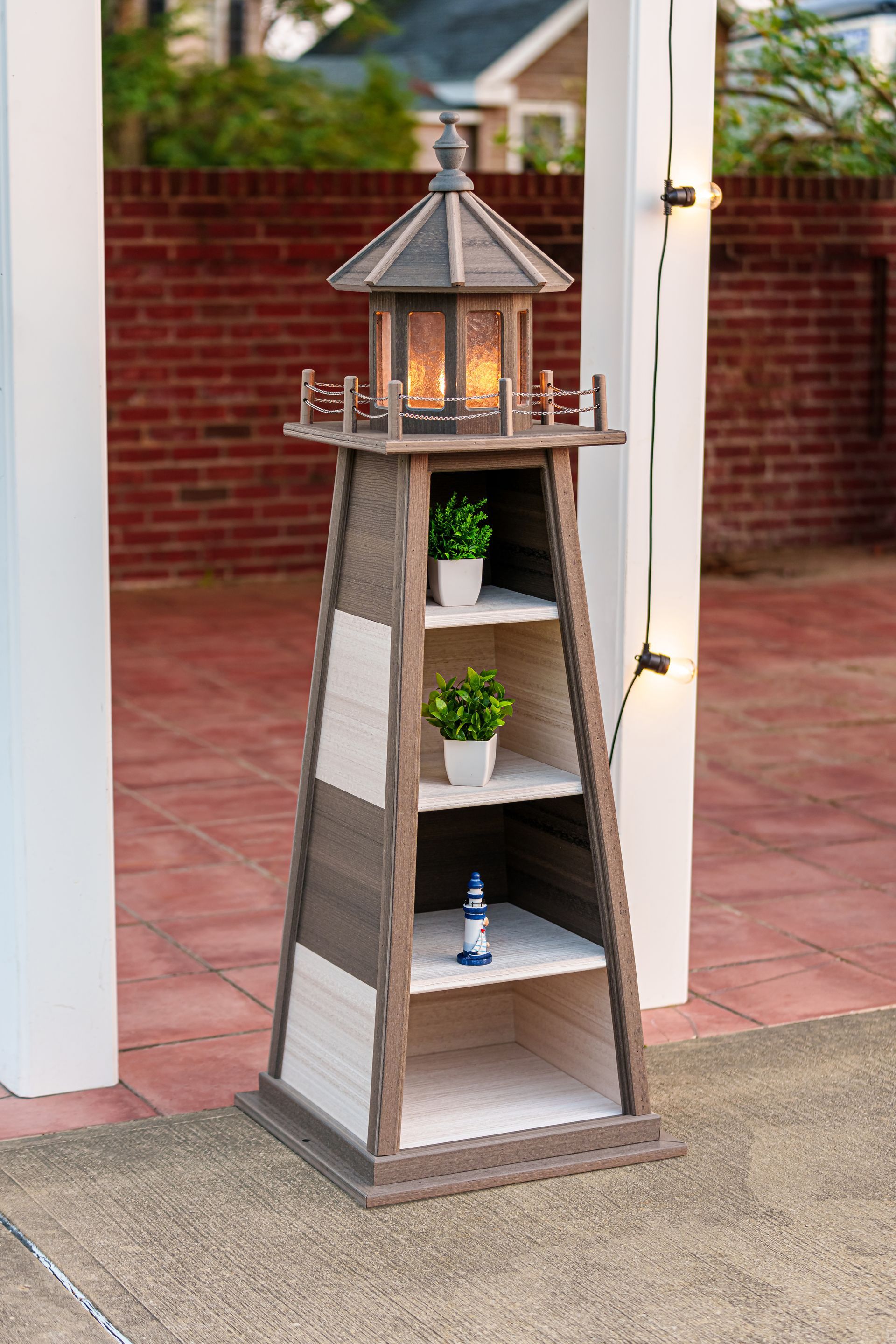 A rustic, wooden lighthouse-shaped shelving unit with a lit top lantern, standing on a patio against a brick wall.
