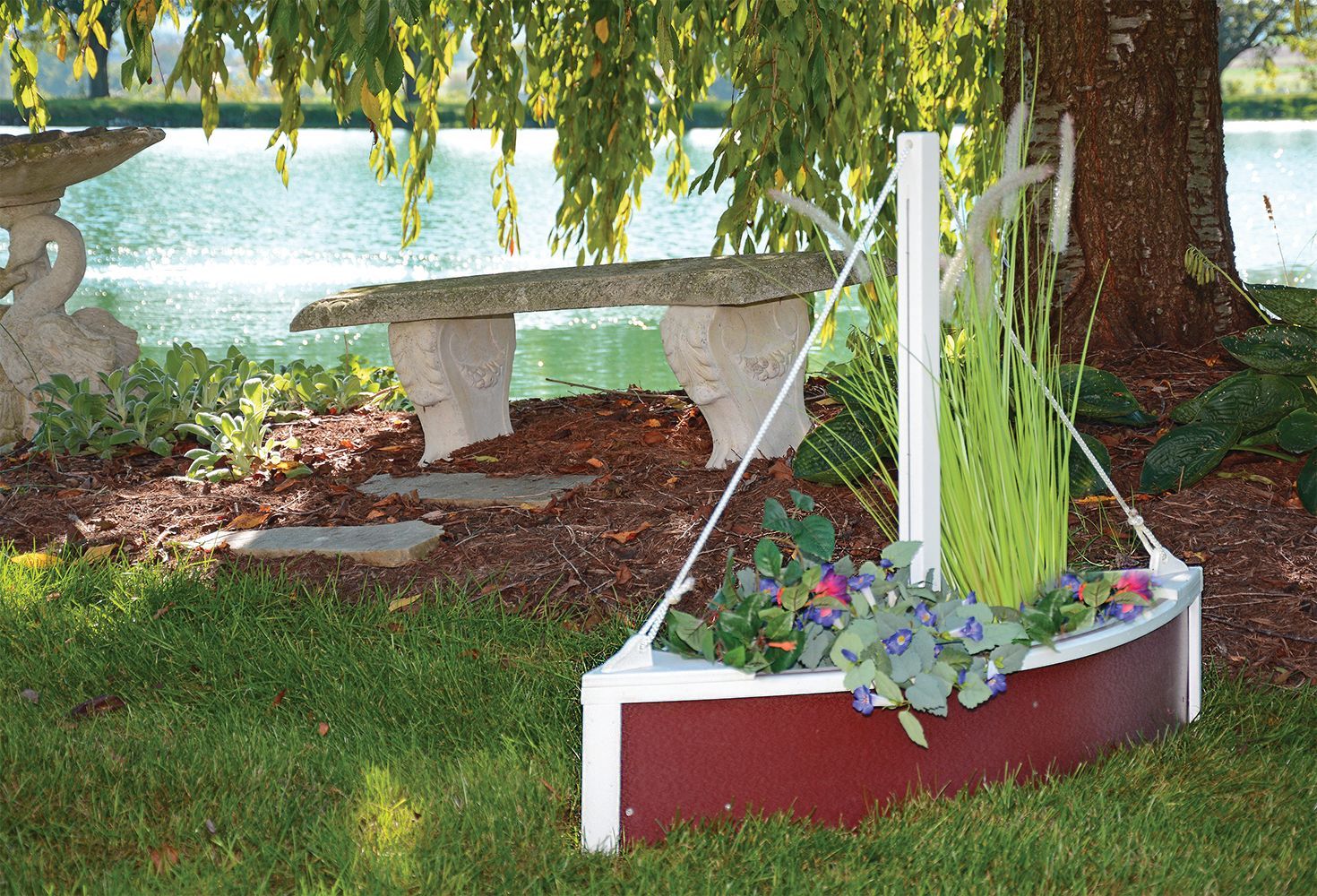 A boat-shaped planter filled with flowers and greenery sits on a lawn, with a pond and bench in the background.