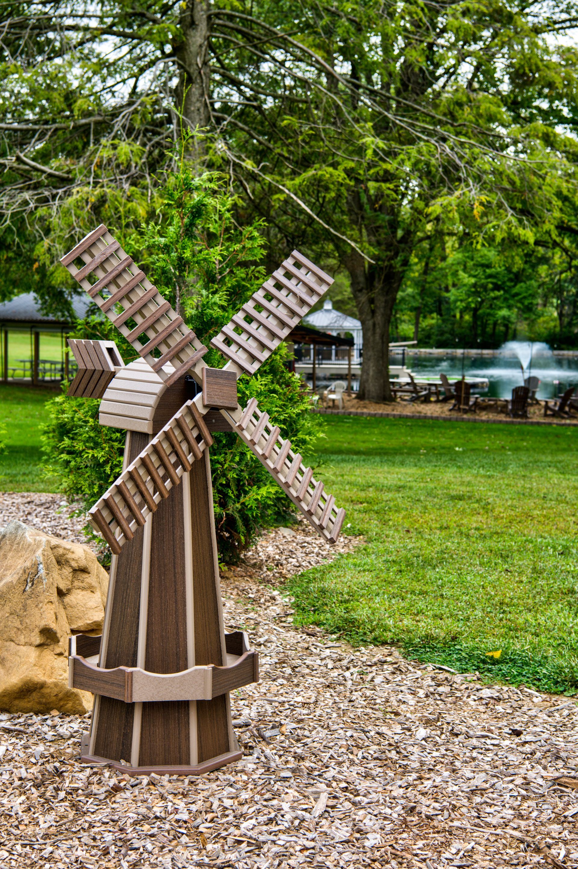 A rustic, brown wooden decorative windmill stands in a gravel garden bed in front of a lush green lawn and trees.