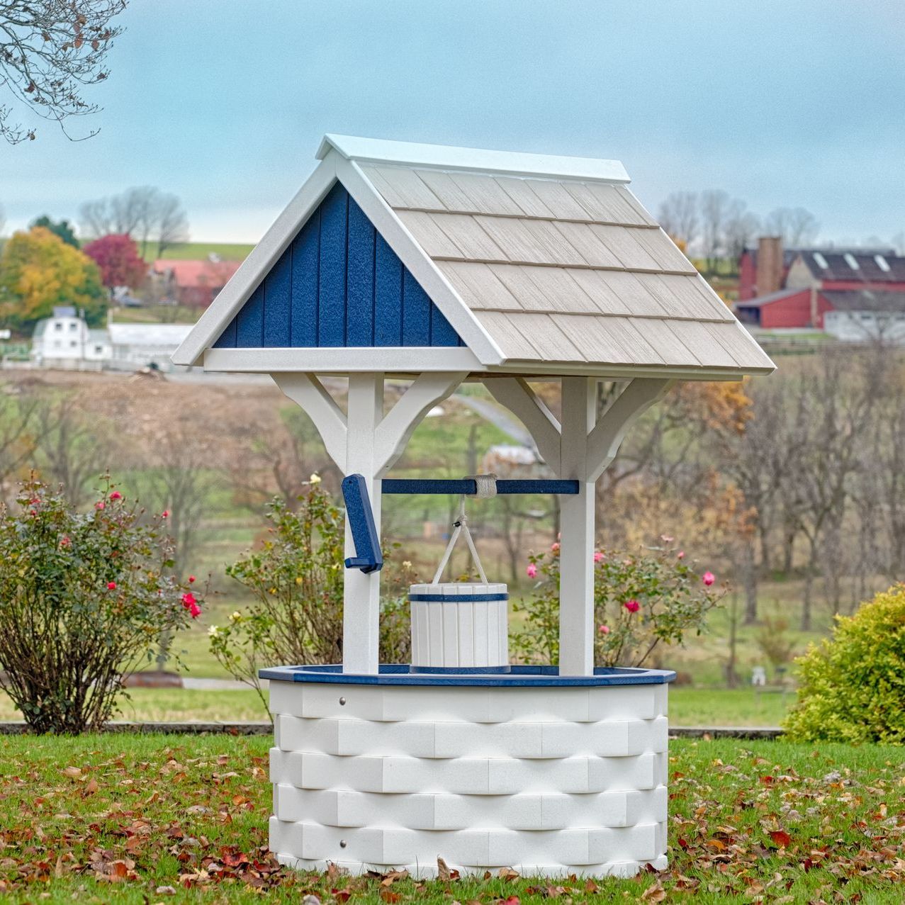 White wishing well with blue and tan accents in a grassy yard, with a rural background.