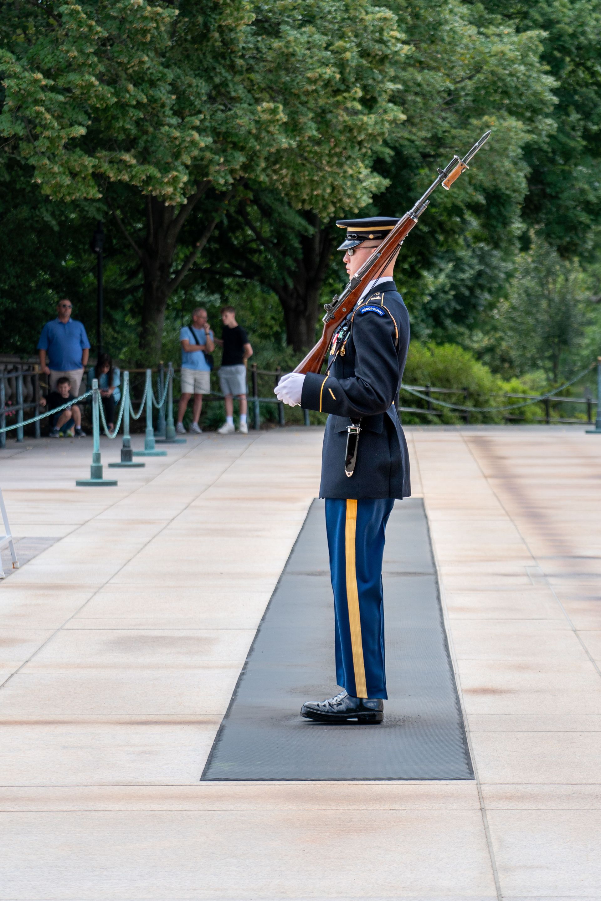 Uniformed guard standing at attention on a walkway, holding a rifle upright, with trees and onlookers in the background