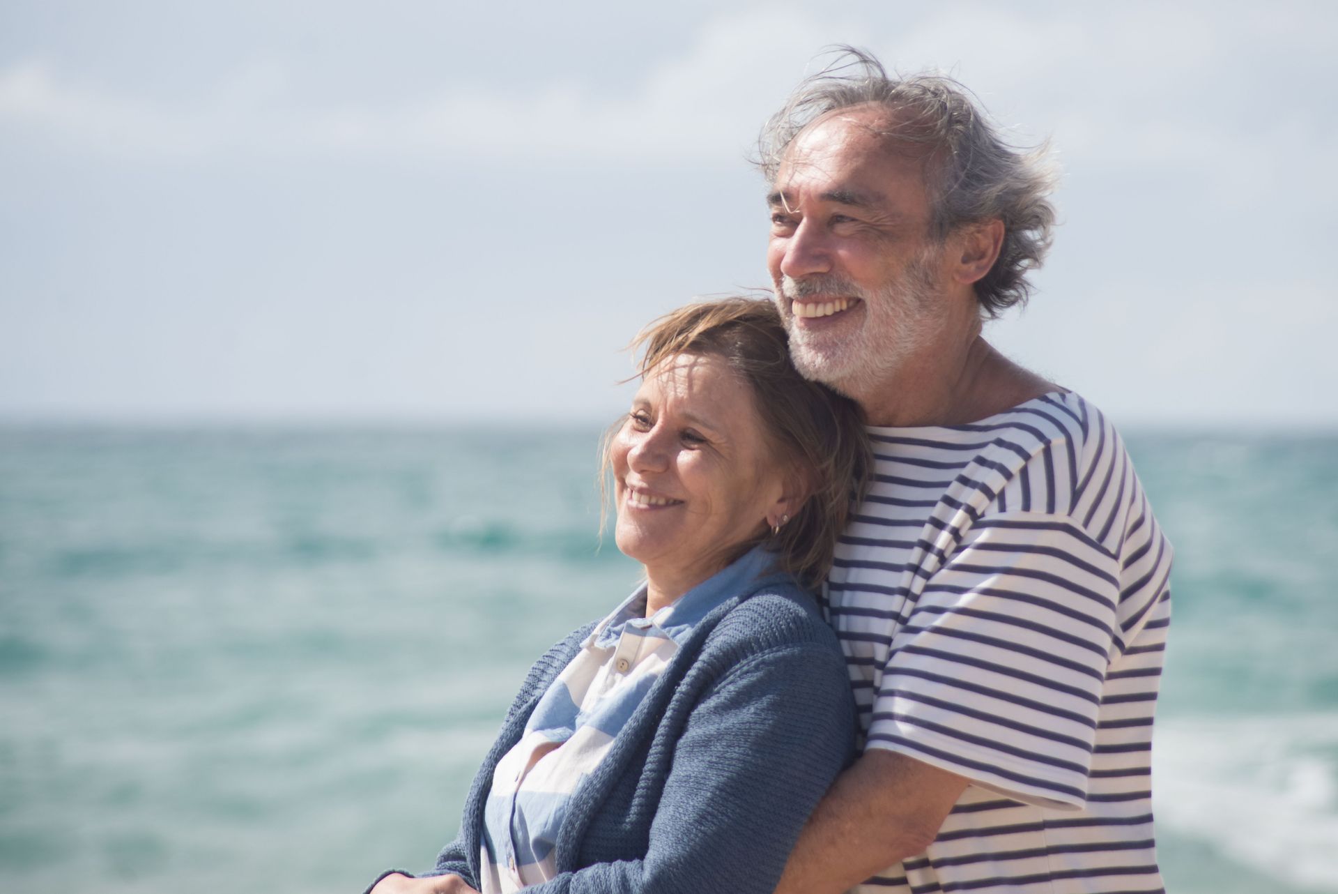 Couple smiling by the sea, with windblown hair and a striped shirt on a sunny day