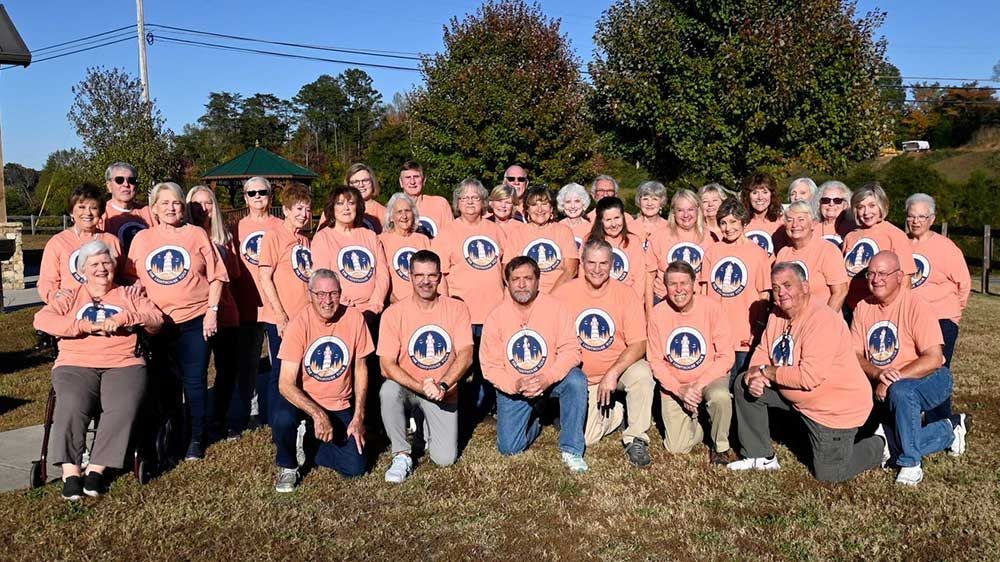 Large group posing outdoors in matching peach shirts with circular logo, standing on grass under a blue sky