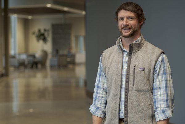 Man smiling in a plaid shirt and tan vest, standing in a bright office lobby.