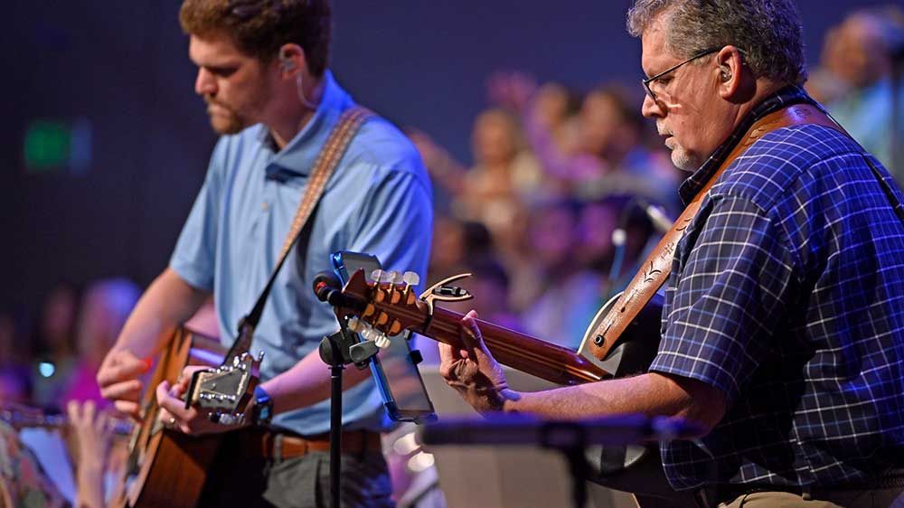 Two musicians playing guitars on stage, one in a blue shirt and one in a plaid shirt