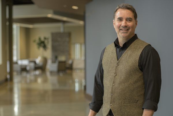 Man smiling in a dark shirt and tan vest standing in a bright lobby