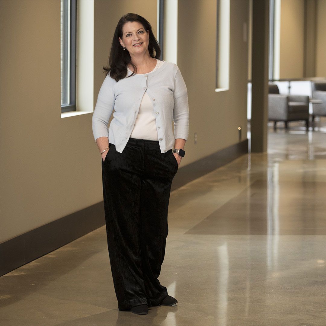 Woman standing in a hallway, wearing a light jacket and dark pants, with hands in her pockets.