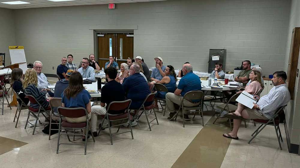 People seated around tables in a meeting room, with a presenter at the front and a wall-mounted screen.