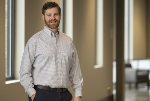 Smiling man in a light checkered shirt stands in an office hallway.