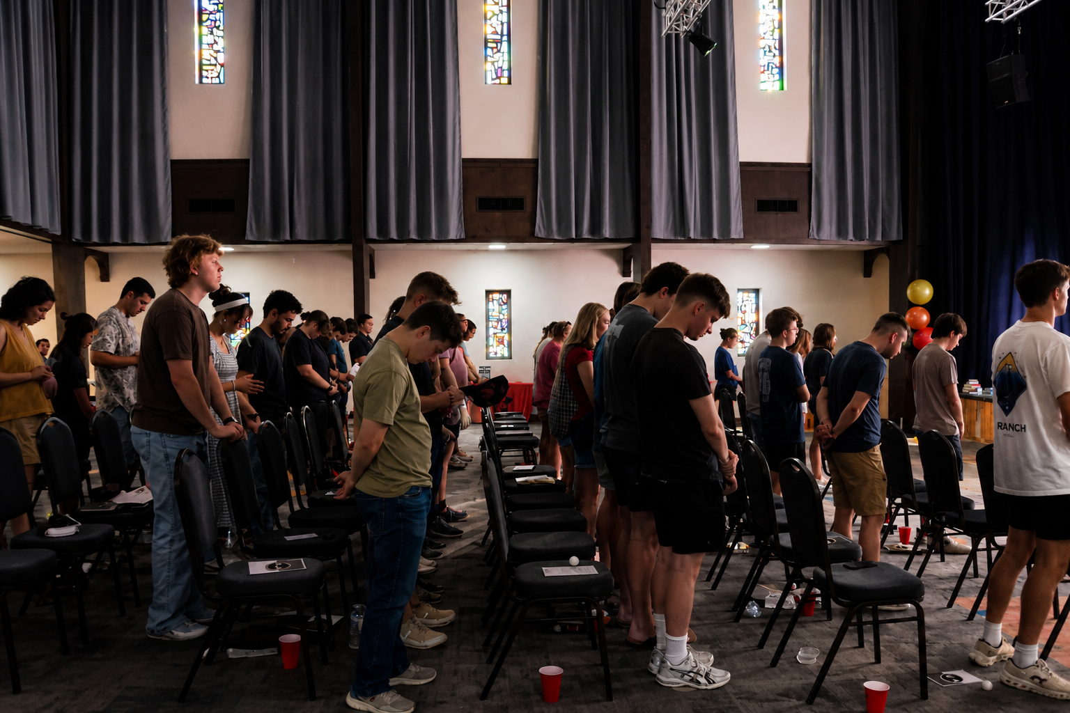 People standing between rows of chairs in a church-like hall with tall stained-glass windows.