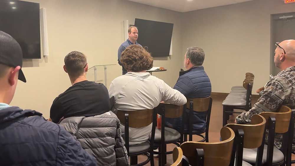Small group seated in a room listening to a man speak near a wall-mounted TV.