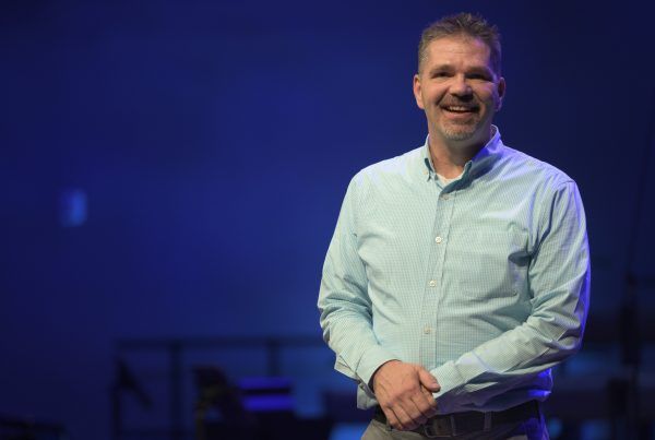 Smiling man in a light blue shirt standing on a blue-lit stage