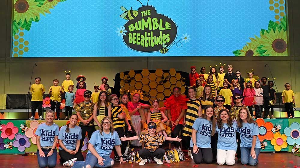 Large group posing on a colorful school stage beneath a “The Bumble Beeline” sign.