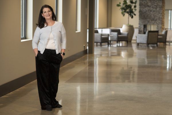Woman standing in a bright hallway, wearing a white jacket and black pants.