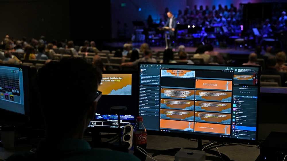 Control room with monitors showing a conference stage and audience under blue lighting.